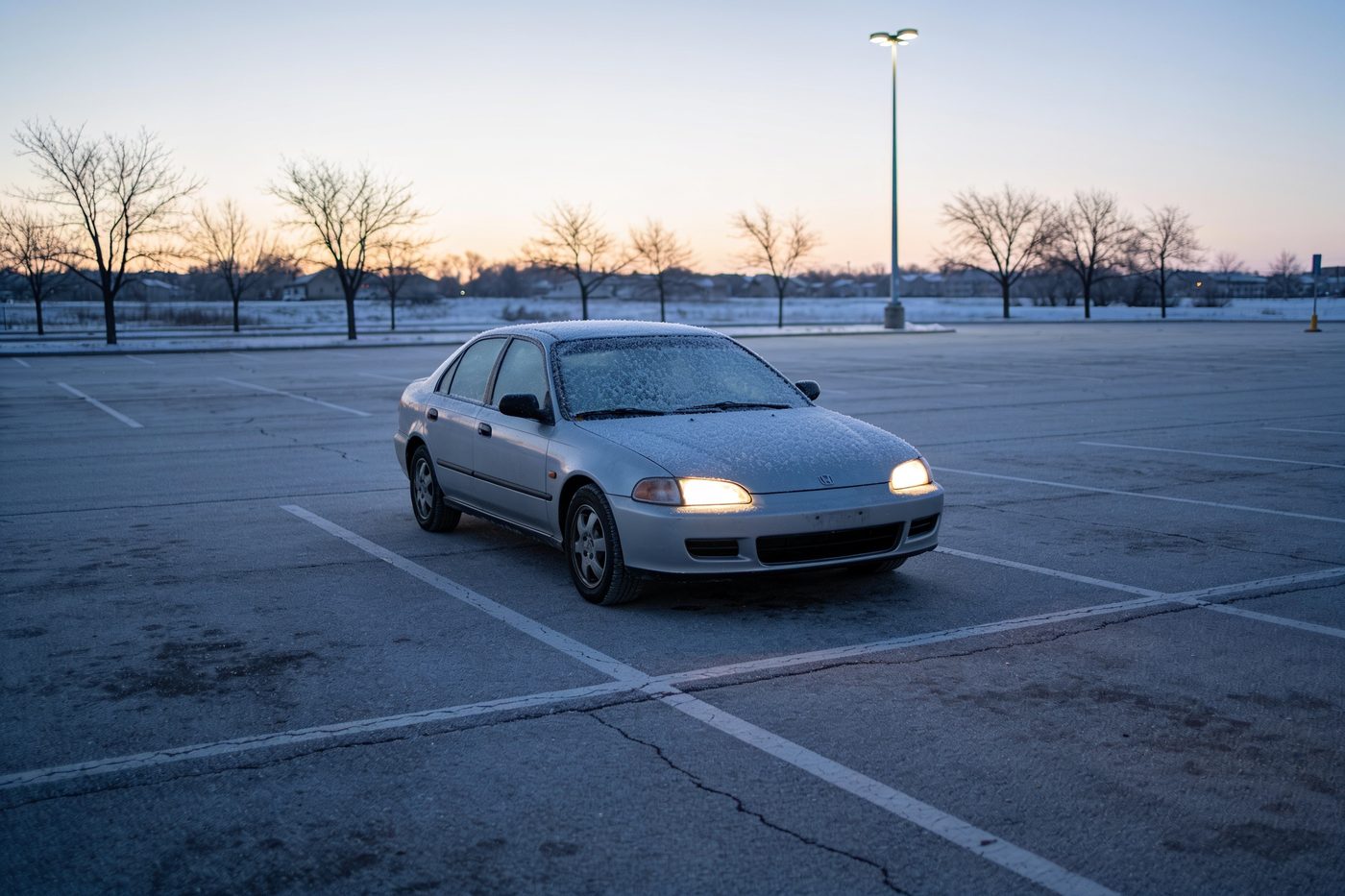 The car alone in an empty snow-edged park and ride lot before seven in the morning, headlights on, frost still across the windscreen and the sun barely up behind bare trees.